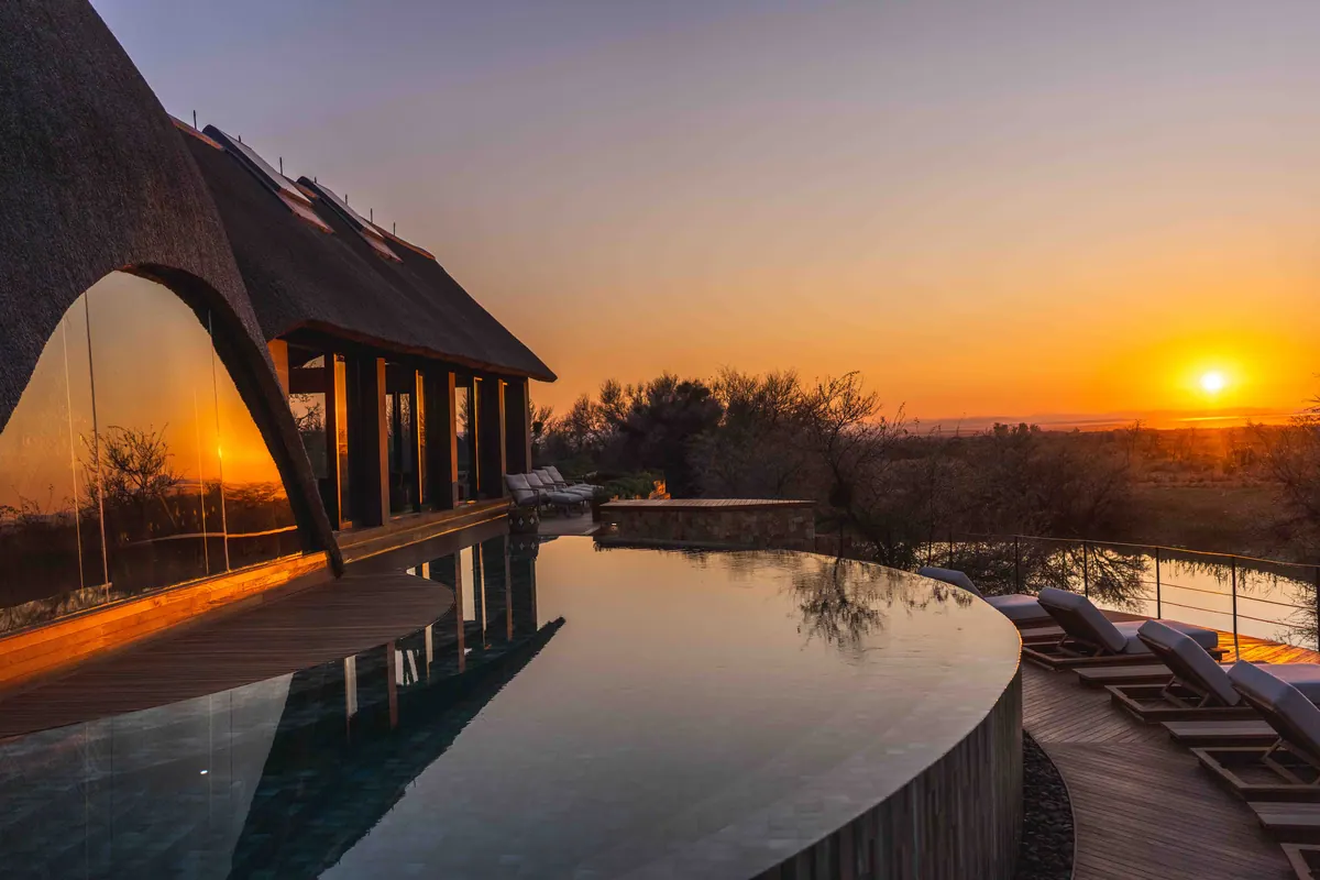 An infinity pool reflecting the sunset beside the thatched lodge